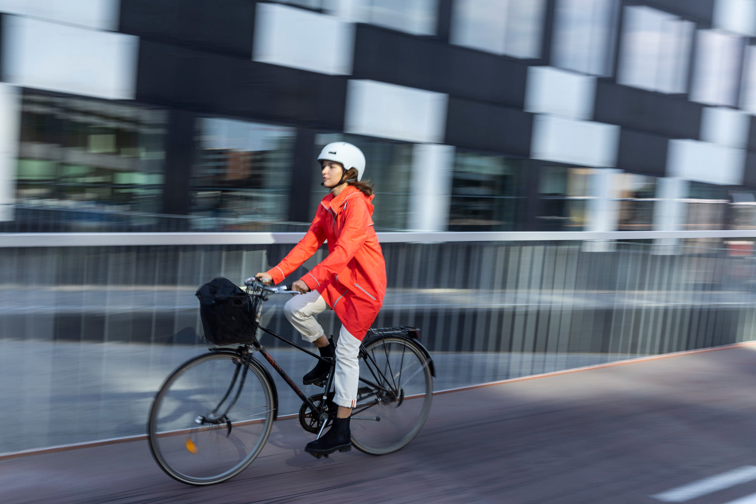 A beautiful model rides a bicycle in a stylish cinnabar red reflective hi vis jacket water resistant fabric with a window pane reflective pattern and reflective piping wears helmet